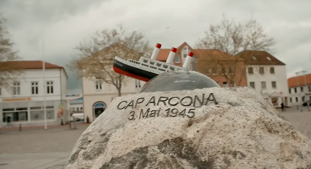 Screenshot aus Video zum Cap-Arcona-Gedenkstein: Zu sehen ist ein Modell des Schiffes, das aus einem rund geschliffenen Teil des Felsstein ragt (einen Untergang symbolisierend), darunter im unbearbeiteten Teil des Felsbrockens eingraviert: »Cap Arcona 3. Mai 1945«