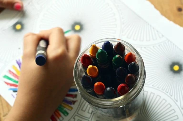 Kinderhand mit blauem Stift im Griff, daneben Glas mit vielen Buntstiften – auf einem Papier mit Mustern zum Ausmalen.