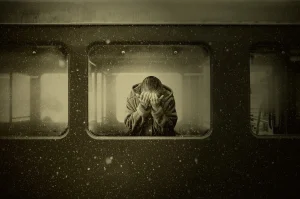 Frau mit Kapuzenpullover im Inneren eines Eisenbahn-Waggons, stehend hinter einer Scheibe, vergräbt ihr Gesicht in ihren Händen. Sie scheint zu trauern. Vor der Scheibe fällt Schnee.
